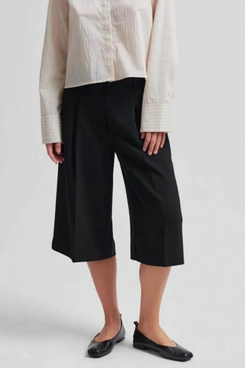 A model wearing black cropped wide-leg pants with a cream-colored textured blouse and black leather ballet flats, shown from the waist down against a light gray studio background.