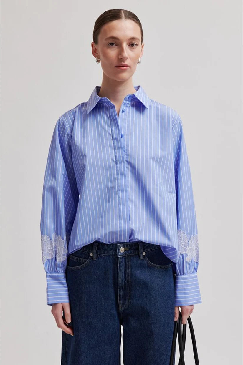 A model wearing a light blue and white vertically striped button-down shirt with white lace embroidery on the cuffs, paired with dark blue jeans, against a neutral gray background.