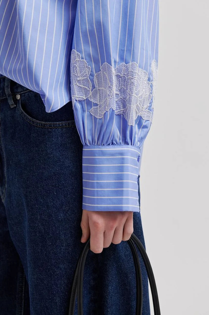 Close-up of a person wearing a light blue and white striped long-sleeve blouse with white floral lace applique on the sleeve, paired with dark blue jeans and holding a black handbag against a light gray background.
