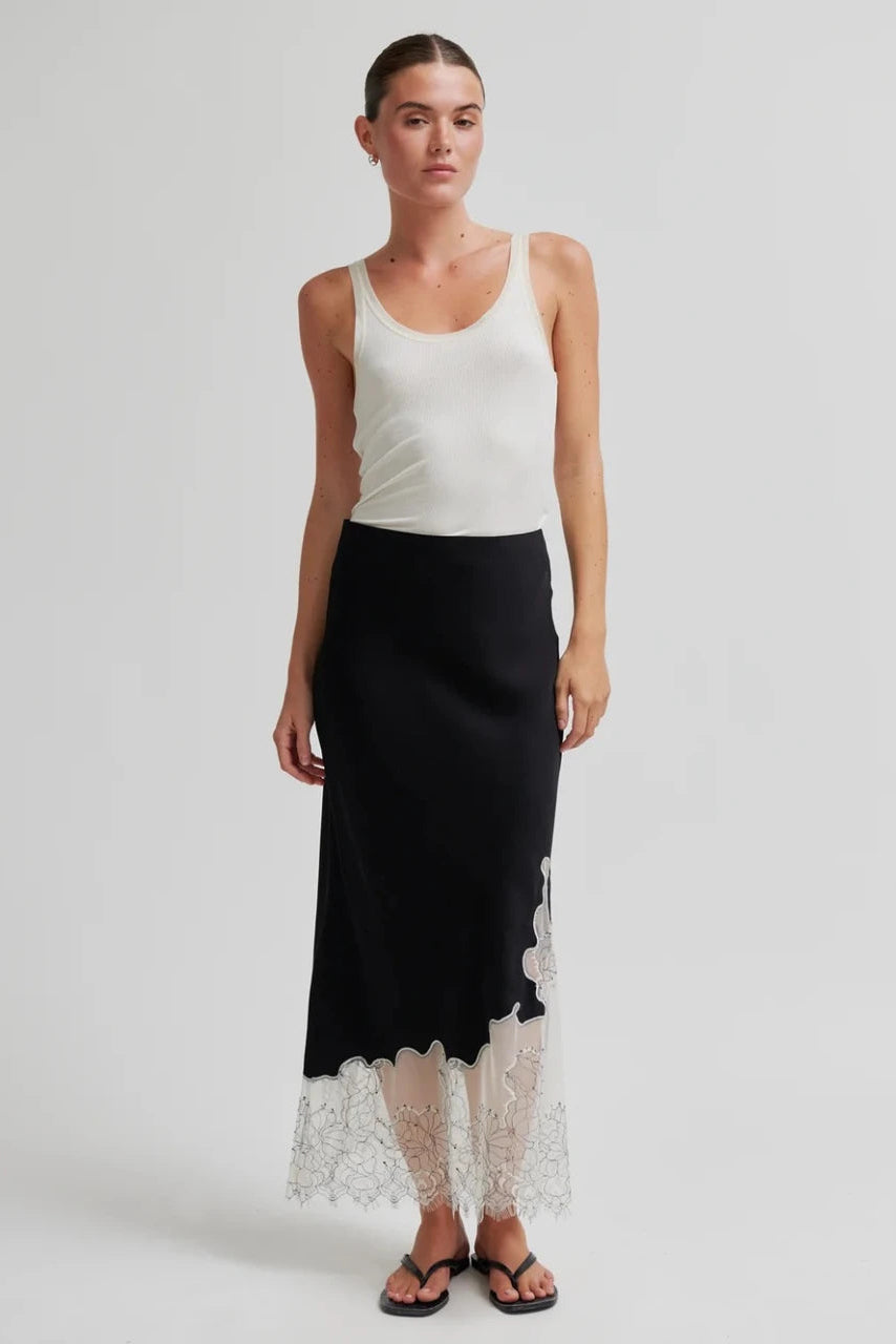 A model wearing a sleeveless white tank top and a black midi skirt with a white lace hem, paired with black flip-flop sandals, against a light gray studio background.