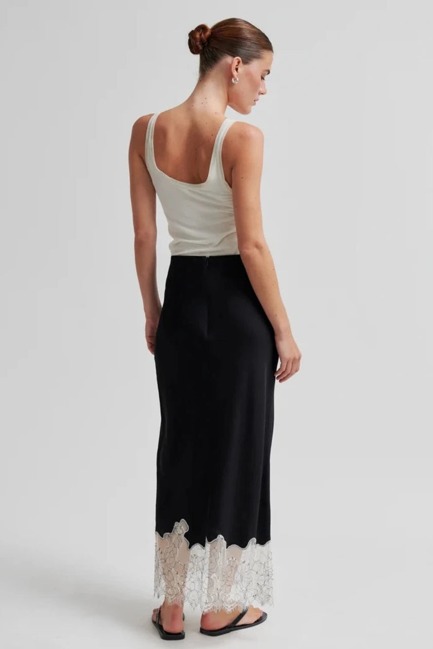 A woman shown from the back wearing a sleeveless black maxi skirt with a white lace hem, paired with a cream tank top, against a light gray studio background.