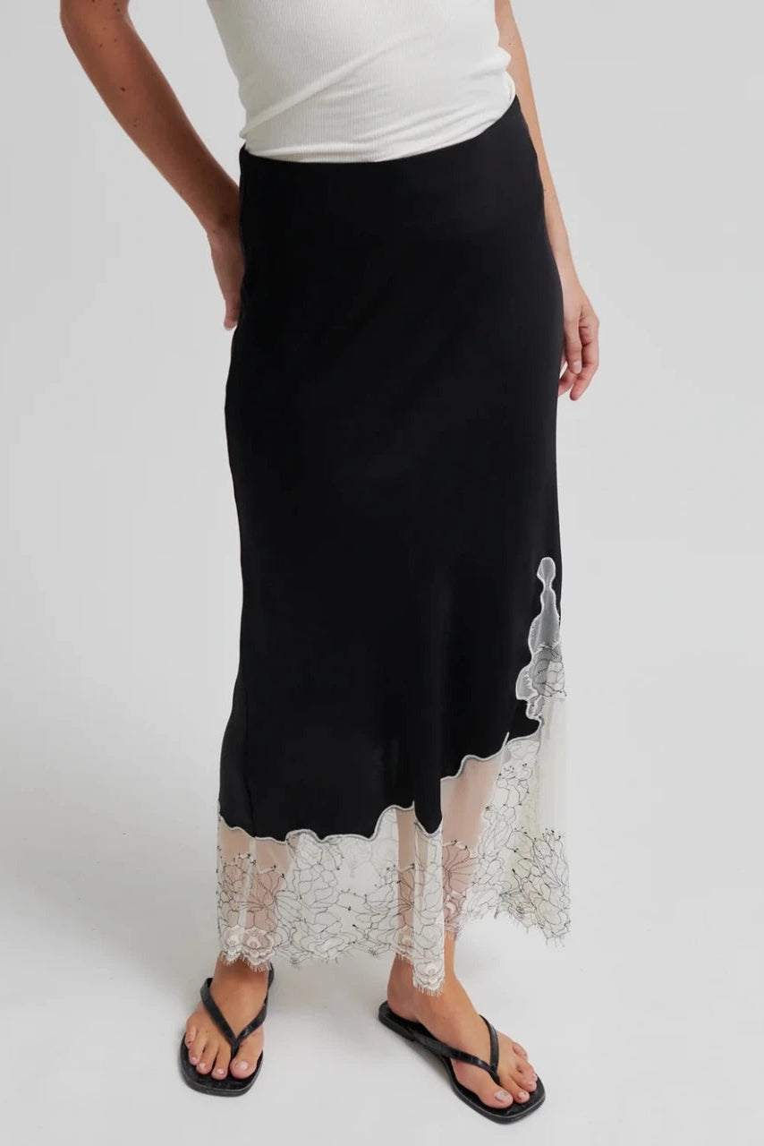 A model wearing a black midi skirt with a sheer white floral lace hem, styled with a white top and black flip-flops against a light gray background.